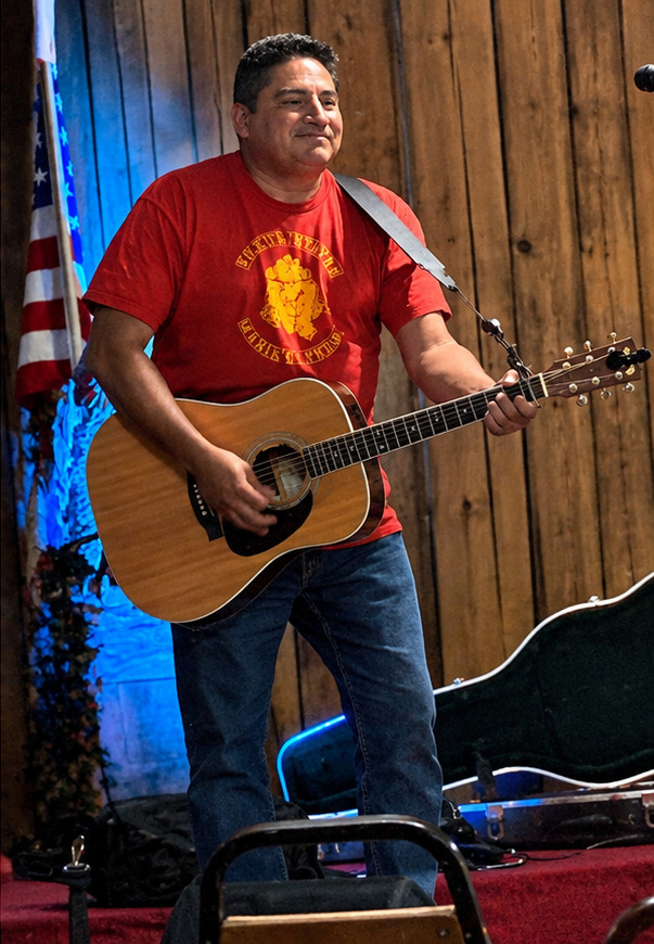 Tom Reyes performing acoustic guitar with an American flag backdrop